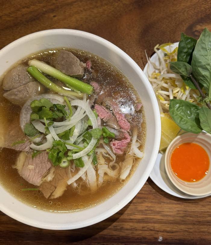 A bowl of phở beside a plate of bean sprouts and herbs