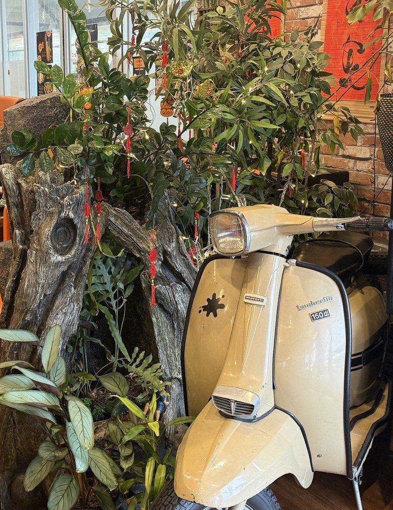 A cream vintage Lambretta scooter parked inside the restaurant against a driftwood-and-plant display, hung with red Tết banners and fairy lights
